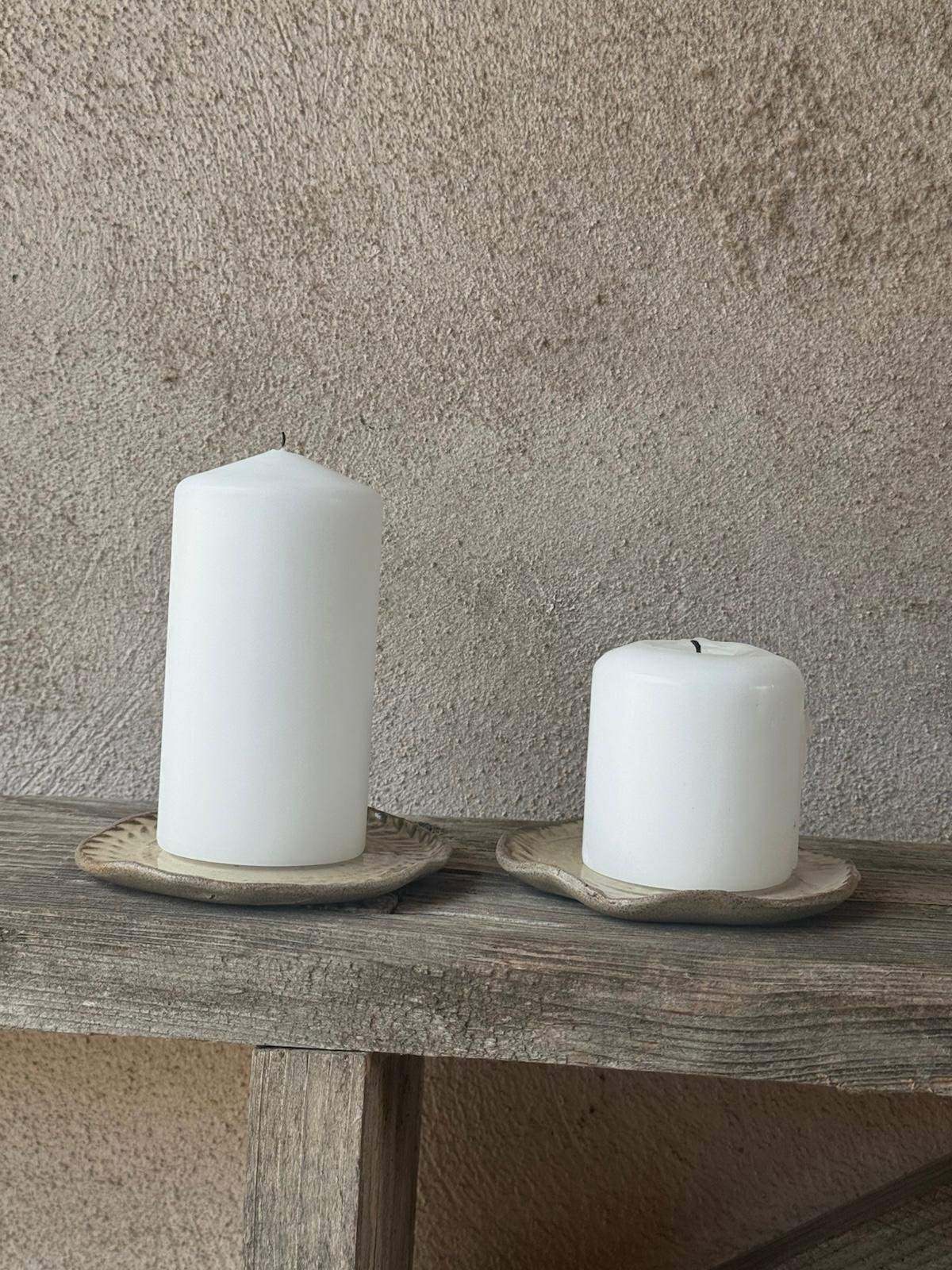 Twee witte stompkaarsen op kleine beige bordjes, op een verweerde houten plank