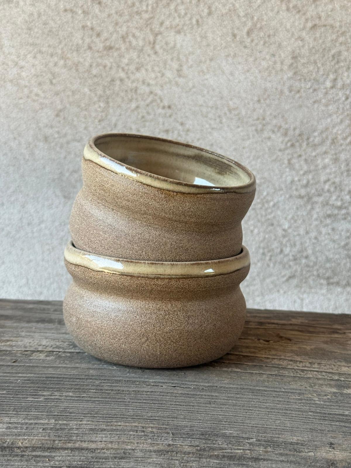 Twee beige bekers met zachte glazuurtinten naast elkaar op een houten plank