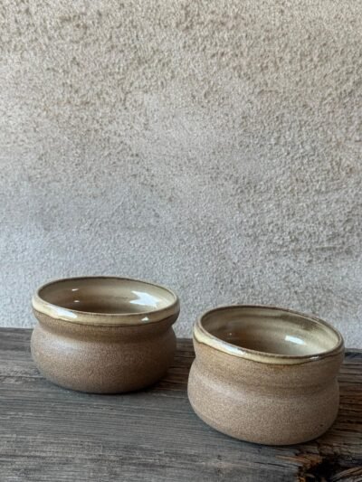 Twee beige bekers met zachte glazuurtinten naast elkaar op een houten plank