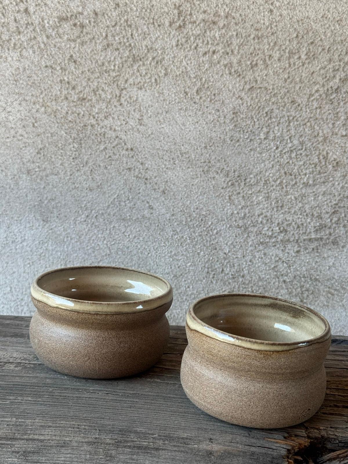 Twee beige bekers met zachte glazuurtinten naast elkaar op een houten plank
