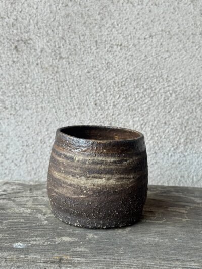 Handgemaakte beker uit donkere, ruwe klei met lichte schakeringen, op een verweerde houten plank