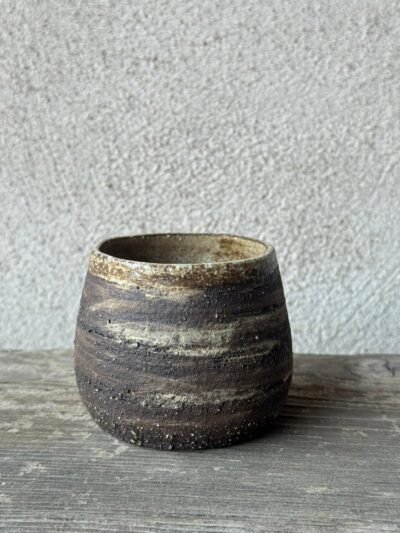 Handgemaakte beker uit donkere, ruwe klei met lichte schakeringen, op een verweerde houten plank