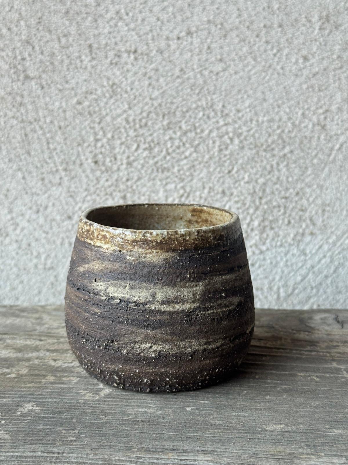 Handgemaakte beker uit donkere, ruwe klei met lichte schakeringen, op een verweerde houten plank