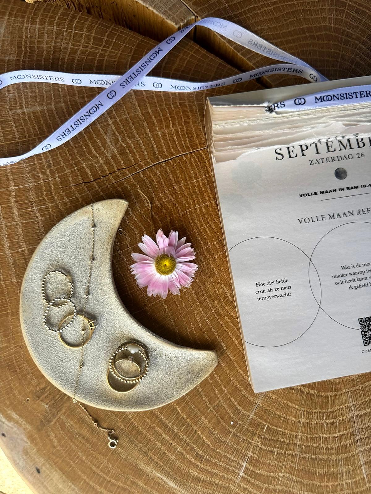 Handgemaakt keramisch maanschaaltje van Holos in de vorm van een halve maan, met ringen en een kettinkje, naast een roze bloem en de Moonsisters-kalender