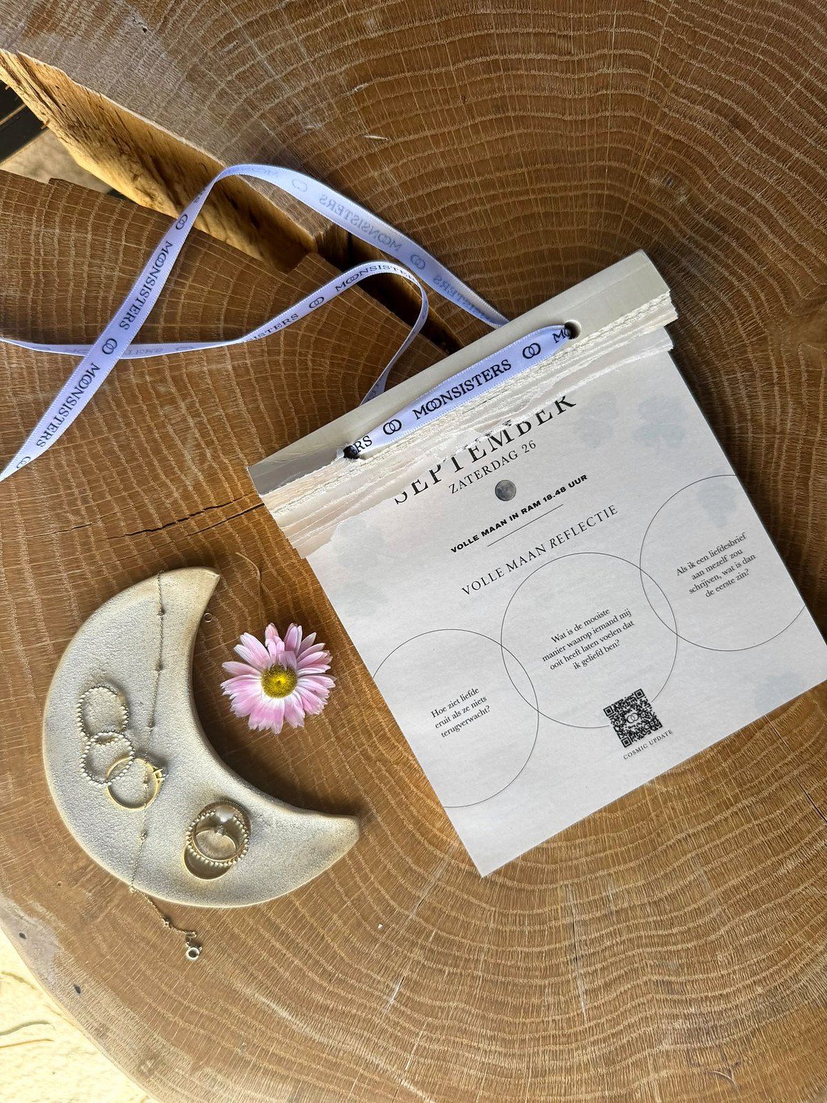 Moon Calendar 2027 van Moonsisters met het lila Moonsisters-lint, naast een handgemaakt keramisch maanschaaltje met ringen en een fijn kettinkje, op een eiken boomschijf