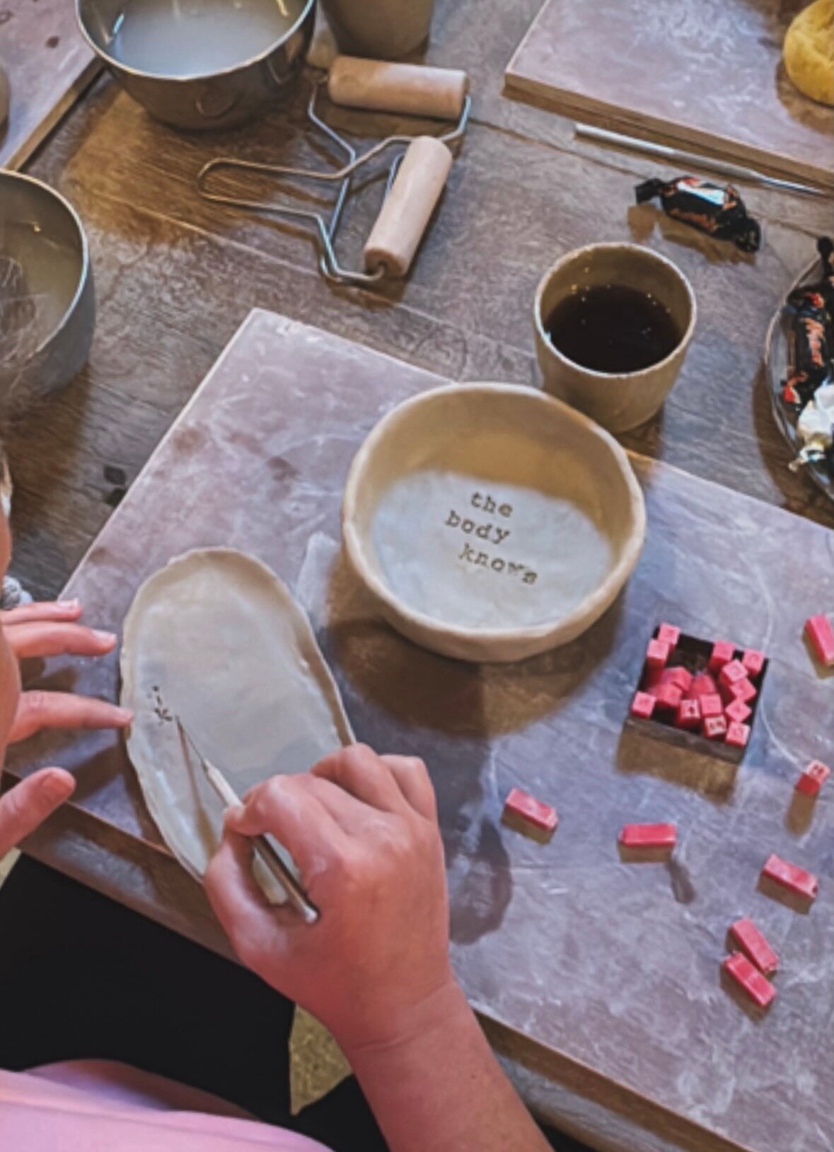 Handen bewerken een schaal van klei aan een werktafel; op een andere schaal staat 'the body knows'.